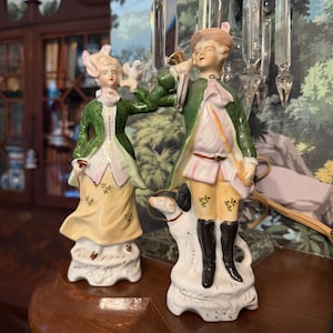 May include: Two porcelain figurines depicting a pair in 18th-century attire. The left figure wears a green jacket and yellow skirt, holding a bird. The right figure wears a green coat, yellow breeches, and black boots, holding a horn with a dog at their feet.