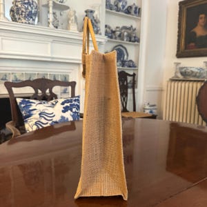 May include: A rectangular, tan burlap tote bag with a light brown handle. The bag is made of a woven material and is standing upright on a dark brown wooden table. The background includes a chair, a fireplace, and shelves with decorative items.