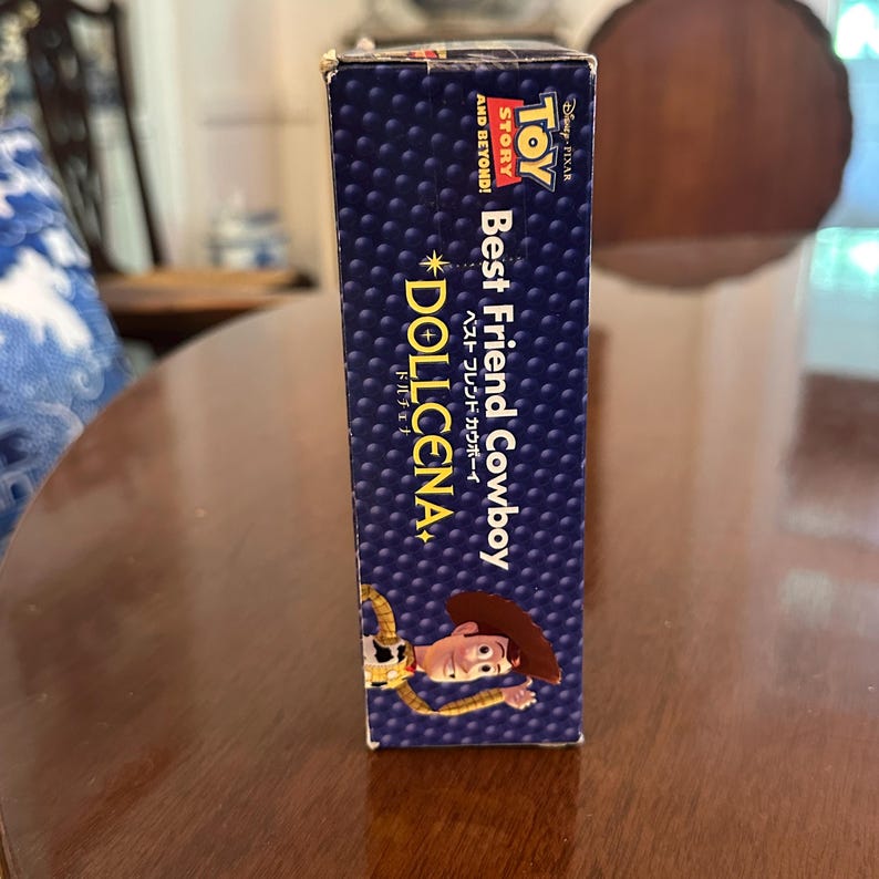 May include: A blue box with the text "Toy Story Best Friend Cowboy" and the character Woody. The box has a dotted pattern and the text "Dollcena" in gold. The box is on a wooden surface.