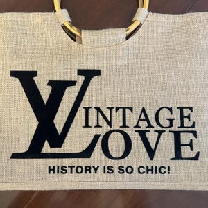 May include: A beige burlap tote bag with a bamboo handle. The bag features the words "VINTAGE LOVE" in black block letters, with the phrase "HISTORY IS SO CHIC!" below.