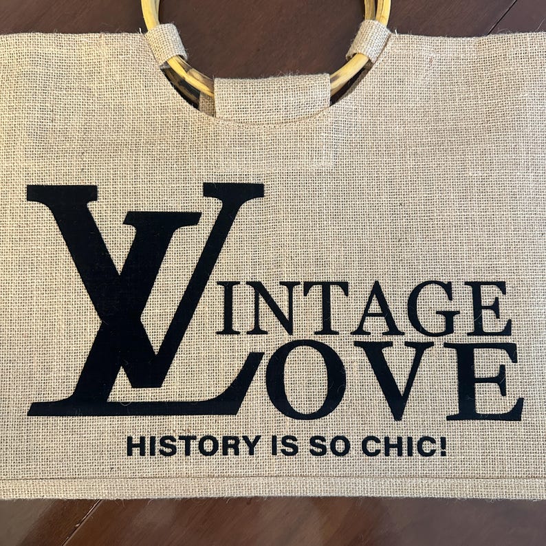 May include: A beige burlap tote bag with a bamboo handle. The bag features the words "VINTAGE LOVE" in black block letters, with the phrase "HISTORY IS SO CHIC!" below.
