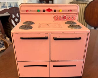 May include: A vintage pink toy stove with four burners, two oven doors, and a control panel with red, green, and yellow accents. The stove has a diamond-shaped emblem and circular dials. The toy is made of metal and has a retro design.