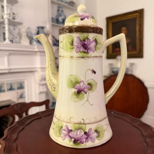May include: A vintage porcelain coffee pot with a cream-colored base, adorned with hand-painted purple violets and green leaves. The pot features a brown rim, a curved spout, and a handle. Gold trim accents the design.