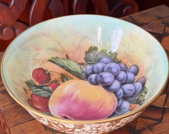 May include: A decorative ceramic bowl with a hand-painted fruit design. The bowl features a peach, grapes, and strawberries. The rim is gold-colored with a decorative pattern. The background has a light yellow and green color.