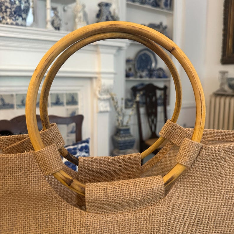 May include: A close-up of a natural burlap tote bag with bamboo handles. The handles are circular and attached to the bag with matching burlap fabric. The bag is a neutral tan color.