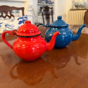 May include: Two small, brightly colored teapots. One is a glossy red, the other is a deep blue. Both have rounded bodies, curved spouts, and small handles. The teapots are sitting on a wooden surface.