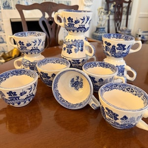 May include: A collection of blue and white porcelain teacups and saucers. The cups and saucers feature a classic blue and white landscape design. Some cups are stacked. The cups and saucers are arranged on a wooden table.