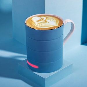 May include: A blue ceramic mug with a white handle sits on a blue charging base. The mug is filled with a light brown latte with a white foam design. The base has a red light.