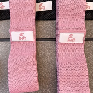 May include: Two pink fabric resistance bands with a white logo that says "H-Fit" and a stylized figure doing a workout.