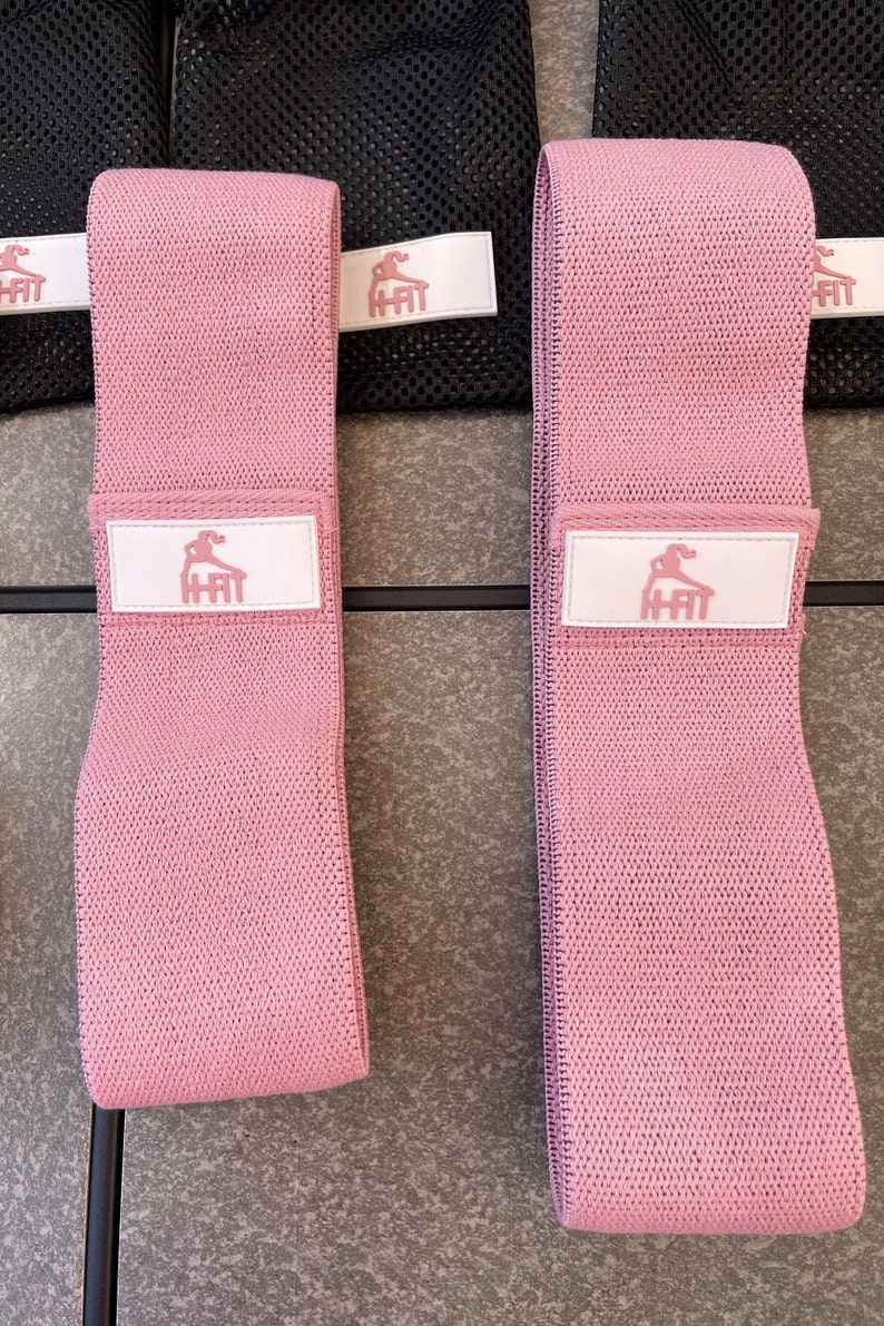 May include: Two pink fabric resistance bands with a white logo that says "H-Fit" and a stylized figure doing a workout.