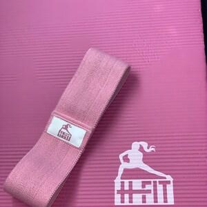 May include: A pink exercise mat with a pink resistance band and a white logo that says "H-Fit" with a silhouette of a woman exercising on top of the text.