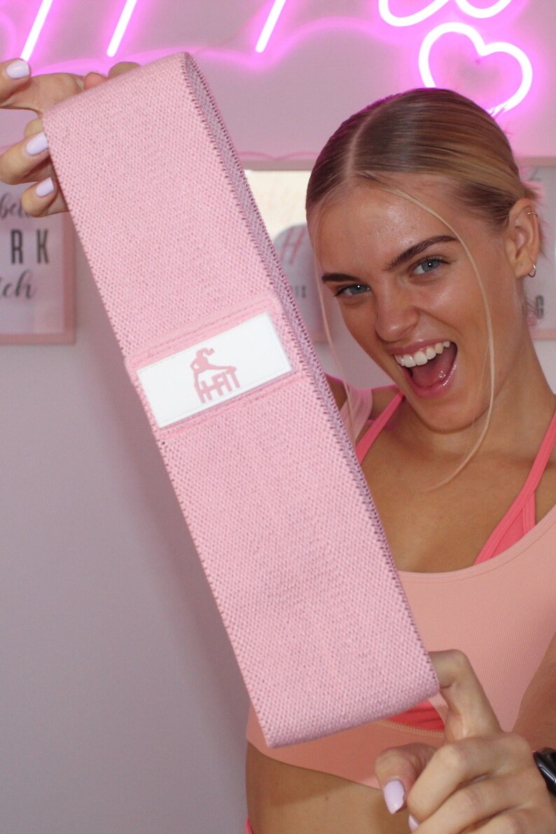 May include: Pink fabric resistance band with a white logo that says "HFit" on it. The band is being held by a person.