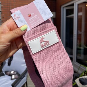 May include: Pink fabric resistance band with a white logo that says "H-Fit" and a small illustration of a woman exercising. The band is rolled up and held in a hand.