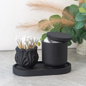 Könnte beinhalten: Ein schwarzes, mattes Badezimmer-Waschtisch-Set. Das Set enthält einen dekorativen Behälter mit Deckel, einen kleinen verzierten Behälter mit Wattestäbchen und ein ovales Tablett. Die Behälter sind aus mattem Material.