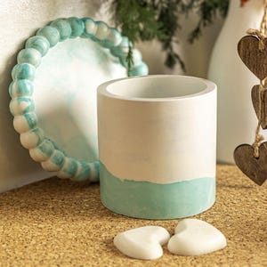 Op de afbeelding: Een cilindrische, wit en blauwgroen geverfde betonnen plantenbak. De plantenbak heeft een wit bovenstuk en een blauwgroen onderstuk. Twee kleine witte hartvormige stenen liggen voor de plantenbak. Een blauwgroene en witte kralen decoratieve cirkel staat achter de plantenbak.