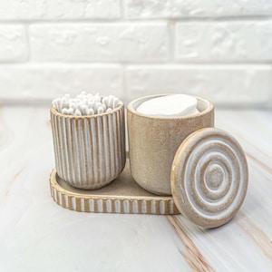 Può includere: Un set di organizzazione per il bagno in ceramica beige. Include un contenitore con cotton fioc, un contenitore con dischetti di cotone e un coperchio. Il set poggia su un vassoio abbinato. I contenitori hanno un design a coste.