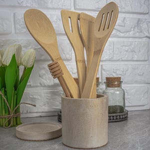 Può includere: Un portautensili in ceramica beige con un set di utensili da cucina in bambù. Gli utensili includono un cucchiaio, una spatola scanalata e un misurino per il miele. Un piccolo coperchio rotondo è davanti al portautensili.
