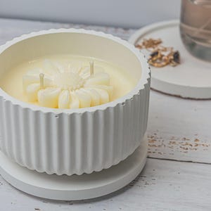 Könnte beinhalten: Ein weißer, geriffelter Kerzenhalter mit passendem Sockel. Die Kerze im Inneren ist hellgelb und hat die Form einer Blume. Die Kerze hat drei Dochte. Der Hintergrund umfasst ein Glas und ein kleines weißes Tablett.