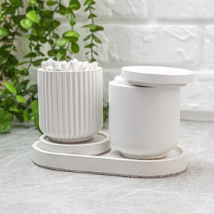 Peut inclure: Un ensemble de deux récipients cylindriques blancs sur un plateau ovale. Un récipient présente une texture nervurée verticale et contient des cotons-tiges. Le second récipient a une surface lisse avec un couvercle blanc assorti posé dessus.