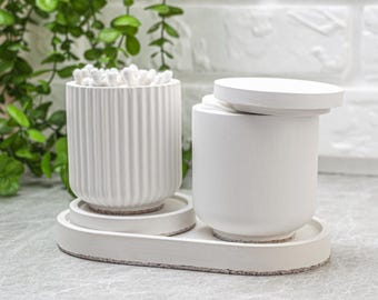 May include: A set of two white cylindrical containers on an oval tray. One container has a vertical ribbed texture and is filled with cotton swabs. The second container has a smooth surface and a matching white lid resting on top of it.