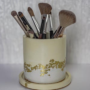 May include: A cream-colored makeup brush holder with gold leaf accents, holding various makeup brushes. The holder sits on a matching round tray with a cork base. The brushes have silver and black handles and brown bristles.
