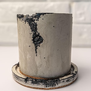 Puede incluir: Una maceta cilíndrica de hormigón con un platillo a juego. La maceta gris tiene una superficie texturizada y una parte superior dentada con detalles en negro. El platillo tiene una base de corcho y un borde pintado en blanco y negro. Diseño moderno y minimalista.