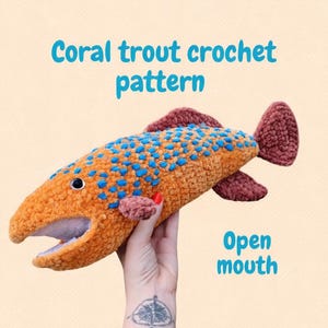 May include: A coral trout crochet pattern is displayed. The plush fish is orange with blue spots and brown fins. The fish has an open mouth. The text "Coral trout crochet pattern" and "Open mouth" are visible.