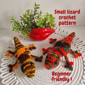 Può includere: Due lucertole all'uncinetto fatte a mano, arancioni, marroni e verdi, con occhi neri, sono esposte su una superficie bianca e strutturata. Una fioriera rossa a forma di labbra contiene piante verdi. Testo: "Small lizard crochet pattern" e "Beginner friendly!"