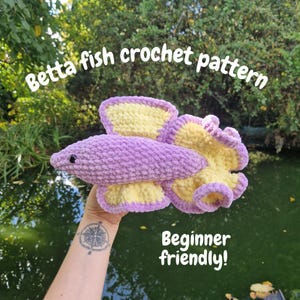 Puede incluir: Un patrón de ganchillo de pez Betta en tonos morados y amarillos. El pez tiene un diseño detallado con aletas grandes y un cuerpo texturizado. El texto en la imagen dice "Betta fish crochet pattern" y "Beginner friendly!"