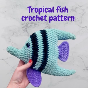 May include: A crocheted tropical fish pattern in shades of teal, black, and purple. The fish has a textured, plush appearance with dark stripes and purple fins. The text "Tropical fish crochet pattern" is displayed in purple.