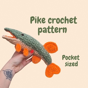 May include: A crocheted fish toy in shades of green, orange, and beige. The text "Pike crochet pattern" and "Pocket sized" are displayed. The fish has orange fins and a detailed design.