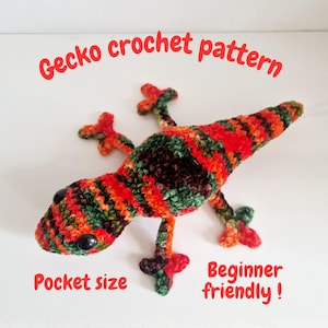 Puede incluir: Un gecko tejido a ganchillo en tonos rojos, naranjas, verdes y marrones. El reptil hecho a mano tiene ojos negros y lleva las inscripciones "Gecko crochet pattern", "Pocket size" y "Beginner friendly!"