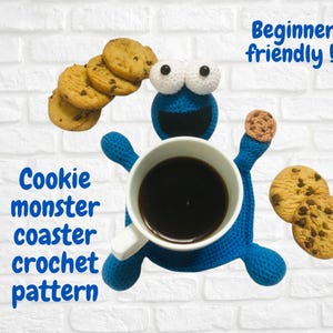 Może przedstawiać: Niebieski szydełkowy podkładka Cookie Monster trzymająca biały kubek wypełniony ciemnym płynem, otoczona ciasteczkami z kawałkami czekolady. Widoczny tekst "Beginner friendly!" i "Cookie monster coaster crochet pattern".