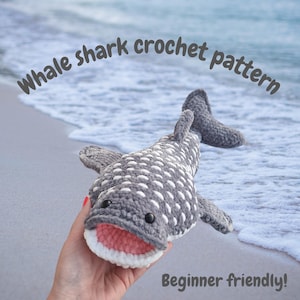 Może przedstawiać: Szydełkowany rekin wielorybi w kolorze szarym i białym z różową paszczą. Widoczny tekst "Whale shark crochet pattern" i "Beginner friendly!". Rekin leży na piaszczystej plaży w pobliżu oceanu.