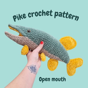 Może przedstawiać: Szydełkowa zabawka w kształcie szczupaka w odcieniach turkusu, beżu i żółci, z otwartą paszczą. Tekst "Pike crochet pattern" i "Open mouth" jest wyświetlany na jasnoniebieskim tle.