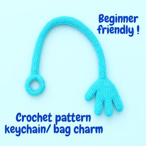 May include: A turquoise crochet keychain or bag charm in a whimsical design. The charm features a loop, a curved handle, and a hand-shaped end. Text on the image reads "Beginner friendly!" and "Crochet pattern keychain/ bag charm."