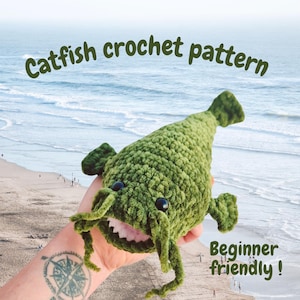 Może przedstawiać: Zielona szydełkowa zabawka w kształcie suma z czarnymi oczami i białymi zębami. Na zdjęciu widnieje napis "Catfish crochet pattern" i "Beginner friendly!". Tło przedstawia plażę i ocean.
