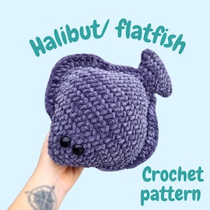 May include: A plush, purple crocheted halibut or flatfish with black button eyes. The text "Halibut/ flatfish" and "Crochet pattern" are written in a teal color on a light blue background. The fish is held by a person.