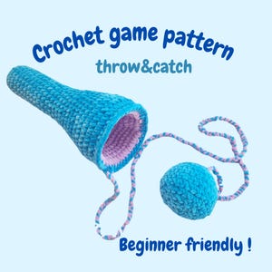 Pode incluir: Um jogo de crochê azul para lançar e pegar, com uma bola presa por uma corda. O interior do lançador é lavanda. O texto na imagem diz "Crochet game pattern throw&catch Beginner friendly!"