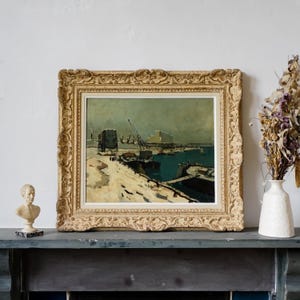 May include: An antique framed painting with a detailed gold-colored ornate frame. The artwork depicts a harbor scene with buildings, cranes, and boats, rendered in muted tones of blue, beige, and gray. The painting is displayed on a dark blue mantel.
