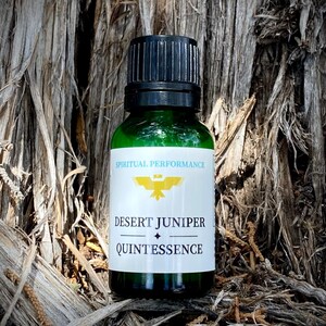 Könnte beinhalten: Eine grüne Glasflasche mit schwarzem Verschluss und einem weißen Etikett mit der Aufschrift "Spiritual Performance Desert Juniper Quintessence". Die Flasche steht auf einer braunen, strukturierten Oberfläche.