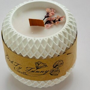 Puede incluir: Vela blanca con una superficie texturizada y una etiqueta marrón que dice "Deh Ty Funny Candle". La vela tiene flores rosas secas en la parte superior.