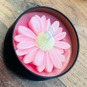 Puede incluir: Una vela de flor rosa en un recipiente de hojalata negro. La flor es una margarita con un centro verde claro y acentos blancos.
