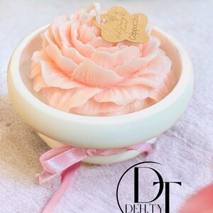 Puede incluir: Una vela rosa en forma de rosa en un cuenco de cerámica blanco con una cinta rosa. La vela tiene una pequeña etiqueta que dice "Deh.Ty Candles" y "Cappuccino".