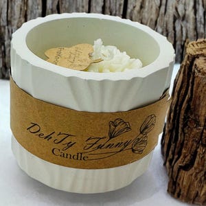Puede incluir: Portavelas de hormigón blanco con una etiqueta de papel marrón que dice "Dek'Ty Funny Candle". La vela es blanca y tiene un diseño floral.