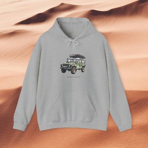 Puede incluir: Una sudadera con capucha gris con un Land Rover Defender verde en la parte delantera. El Land Rover tiene un portaequipajes en el techo y está conduciendo por un paisaje desértico. El texto "RUGGED ELEGANCE" está impreso debajo del vehículo.