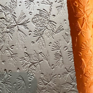 May include: A close-up of a textured, orange rolling pin with a pine cone and pine needle design. The rolling pin is being used to create an imprint on a light grey surface.