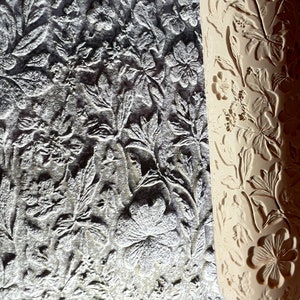 以下が含まれることがあります： 花柄のローラーのクローズアップ。ローラーは木でできており、薄い茶色をしています。デザインはローラーにエンボス加工されており、さまざまな花や葉が描かれています。ローラーは、表面に模様をつけるために使用されています。