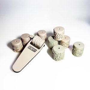 May include: A set of eight beige and green textured rollers with intricate Celtic knot designs. The rollers are used for creating patterns on clay or other materials. The set includes a handheld roller with the word "Potter" printed on it.
