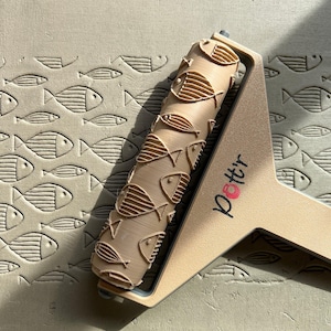 May include: A beige pottery roller with a fish pattern, creating an embossed design on a clay surface. The roller is held by a beige handle with the word "Pottr" printed on it in black and pink.